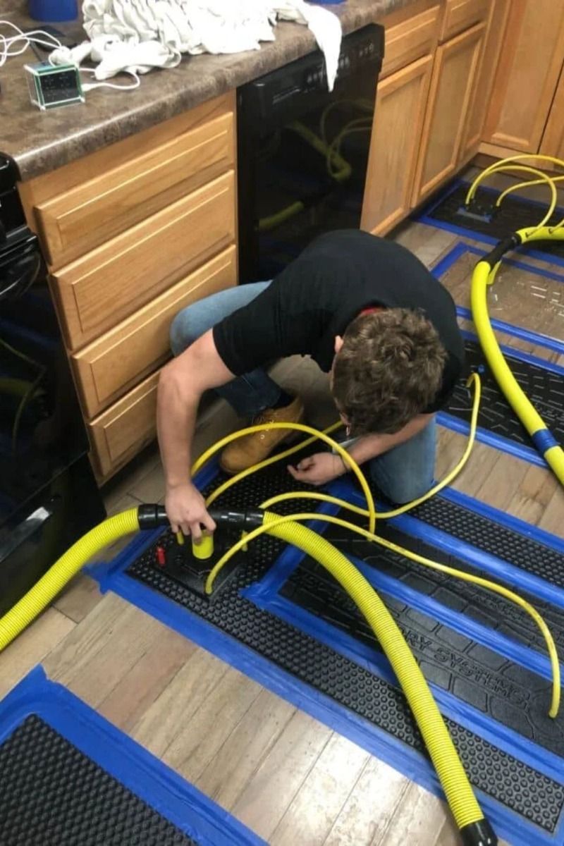 Person kneeling on the kitchen floor, organizing yellow hoses around blue mats and cabinetry.