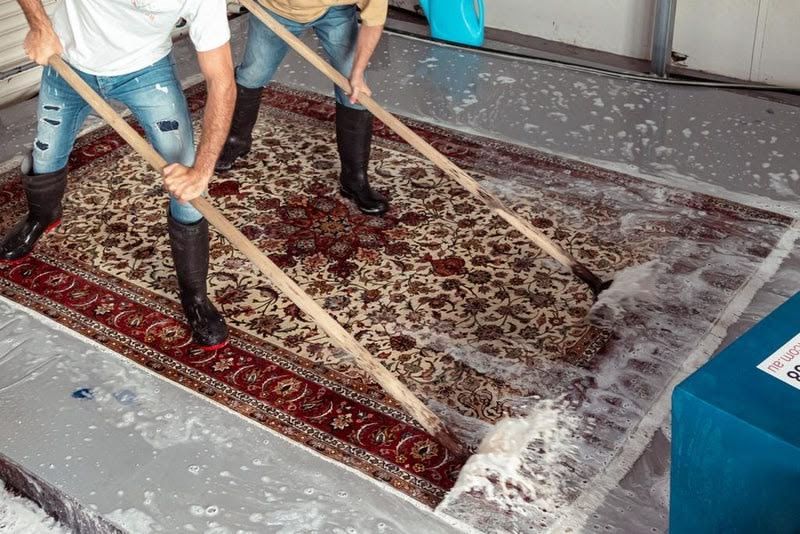 Two people cleaning a patterned rug with brooms on a tiled floor