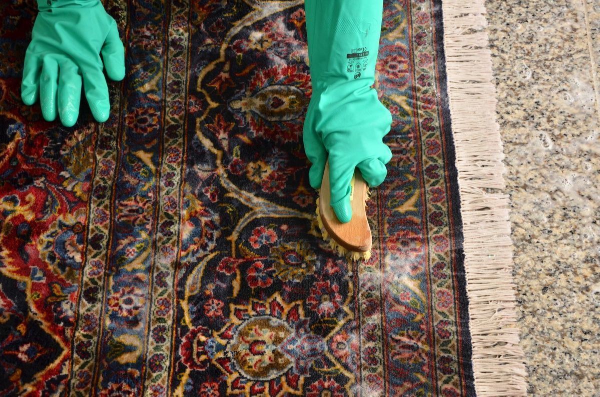 Gloved hands cleaning a patterned rug with a sponge on a speckled floor