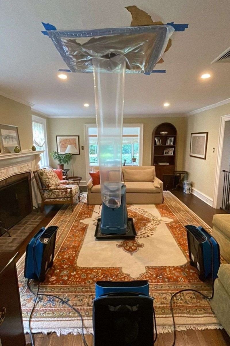 Living room with a ceiling repair setup: plastic sheeting taped overhead and fans on the floor