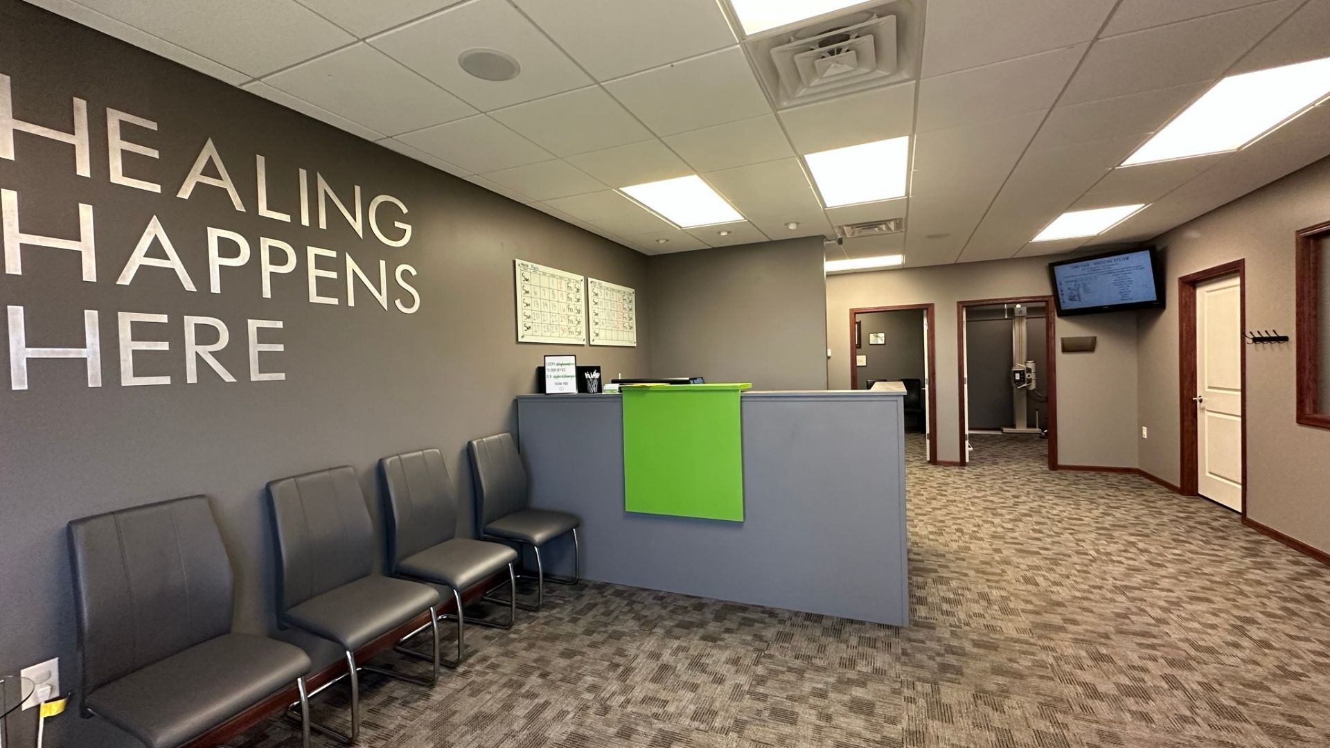 Empty clinic waiting room with gray chairs, reception desk, and wall text reading “HEALING HAPPENS HERE”