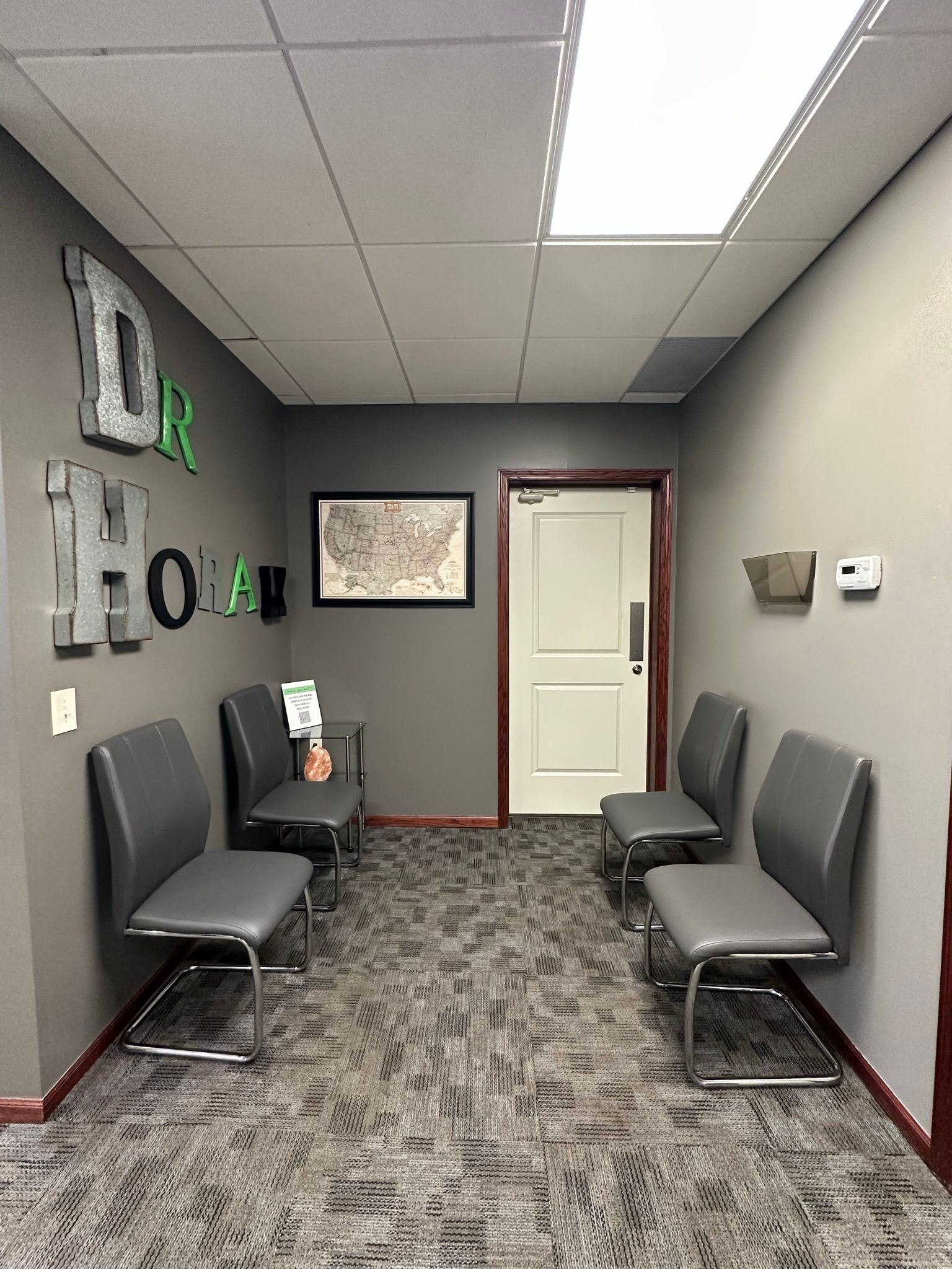 Small waiting room with gray chairs, patterned carpet, white door, and wall decor in muted tones.