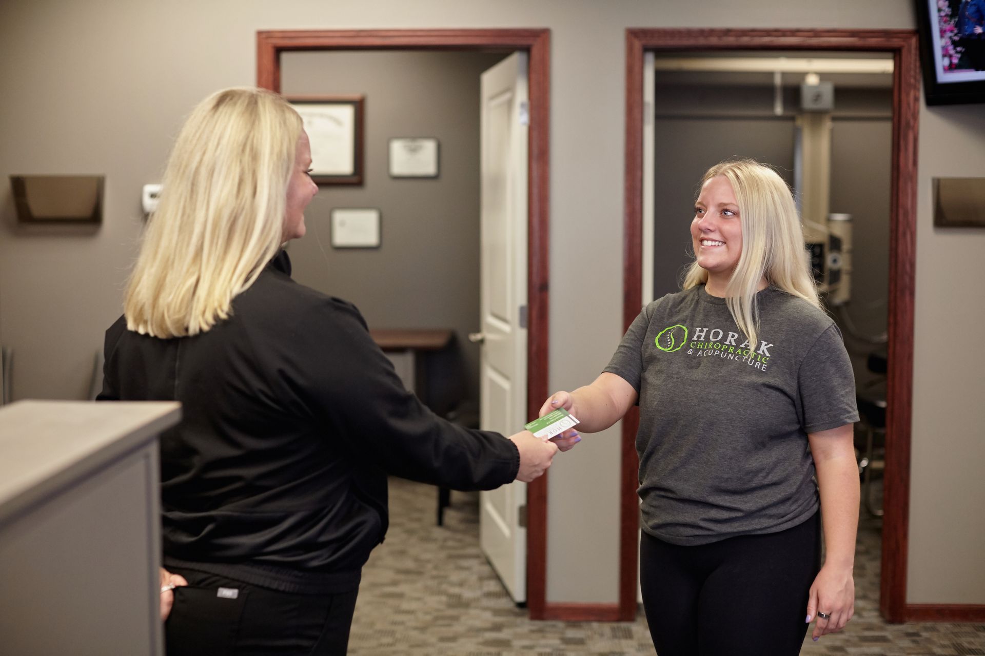 Two women shaking hands in an office hallway, smiling at each other.