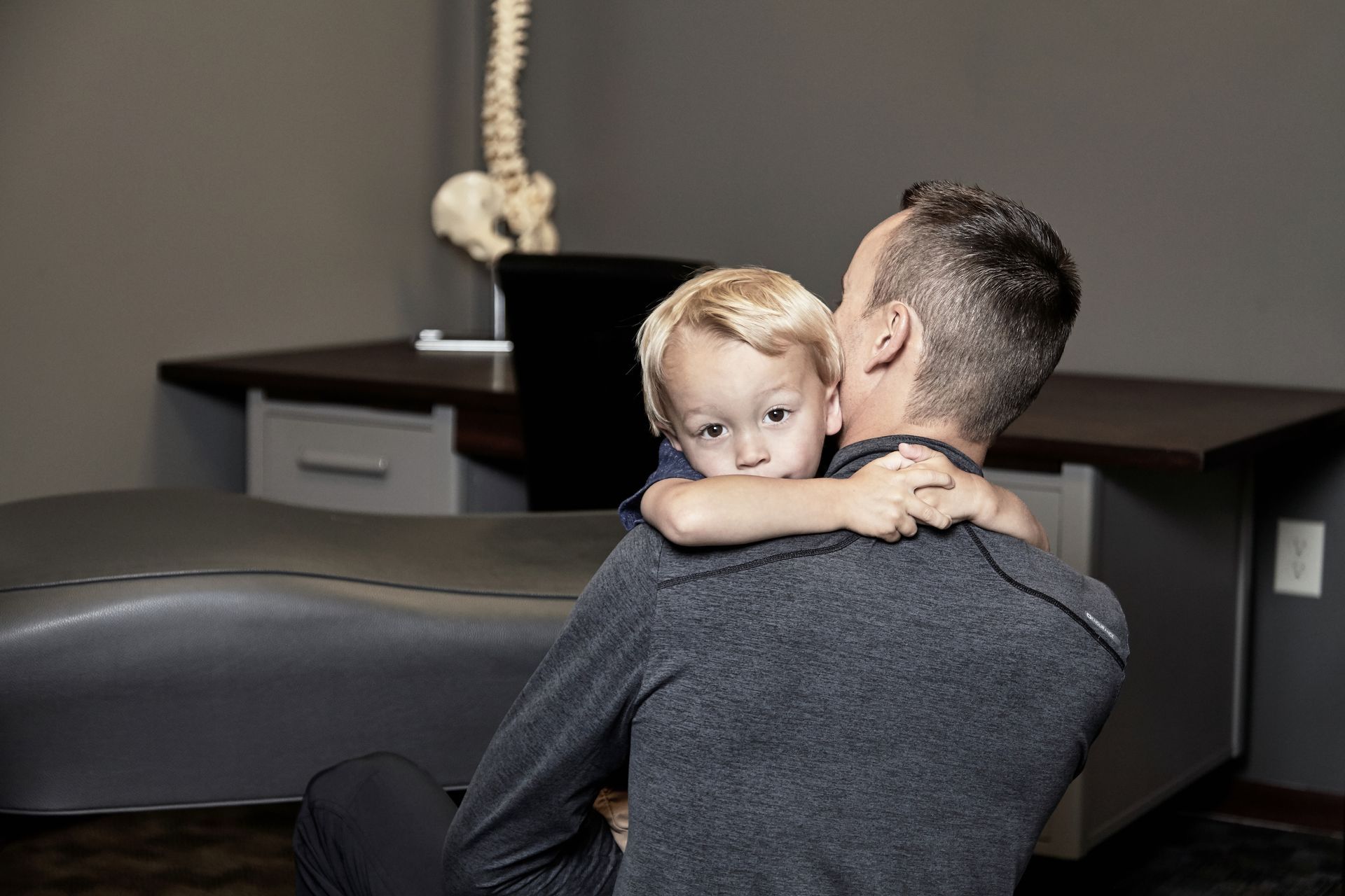 Man holding a child who clings to his shoulder in a dim room with a dark couch and rope in the background