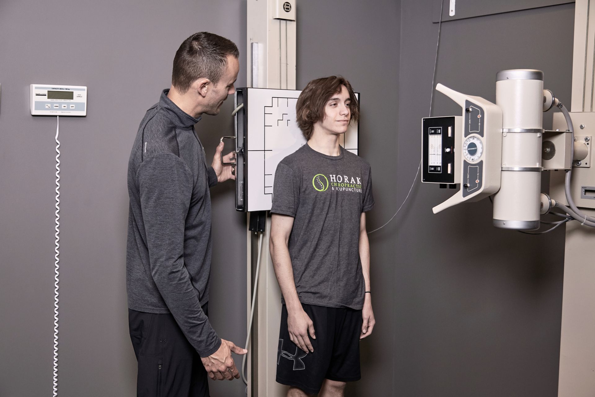 Two people stand beside a medical X-ray machine in a clinic room.