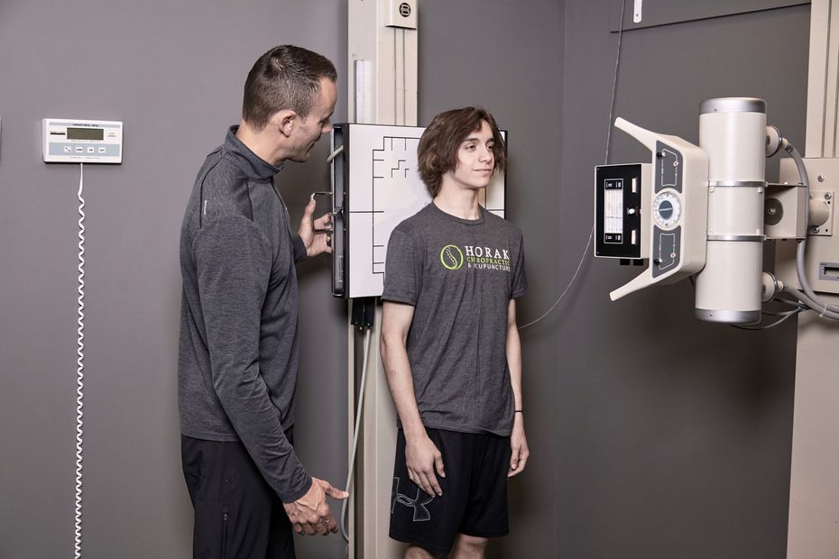 Two people stand beside a medical X-ray machine in a clinic room.