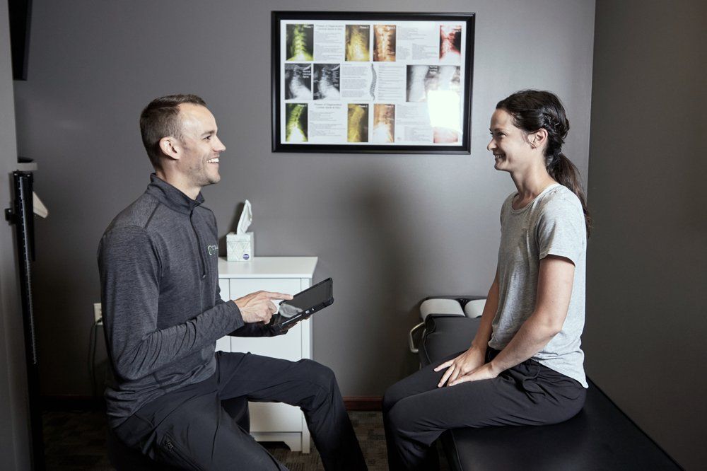 Two people seated facing each other in a clinic room, one holding a device and a chart on the wall behind them.