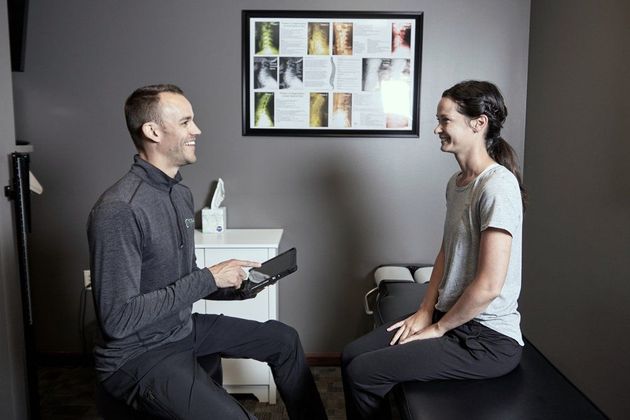 Two people seated facing each other in a clinic room, one holding a device and a chart on the wall behind them.