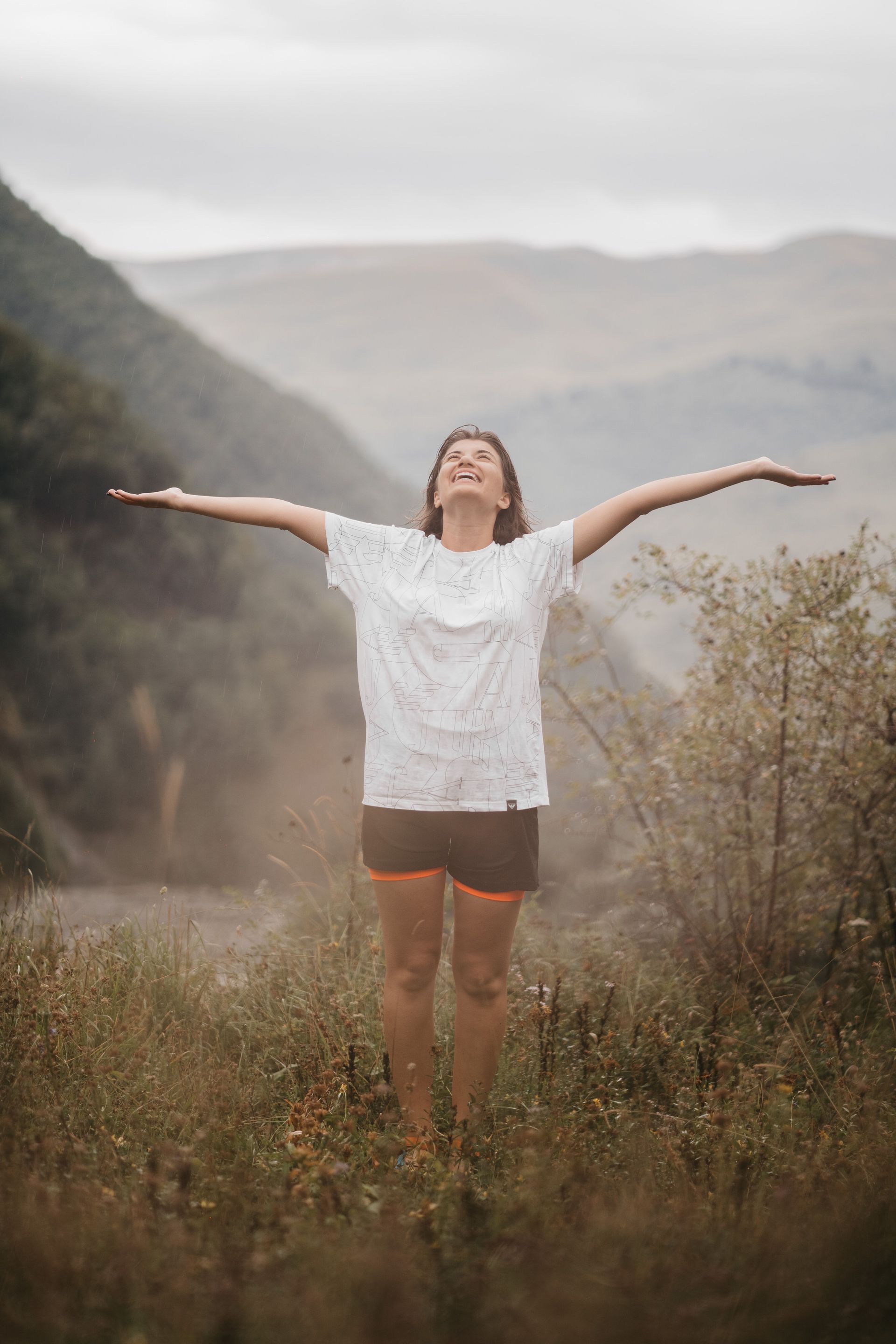 Person standing in a grassy valley with arms outstretched, smiling under a cloudy sky