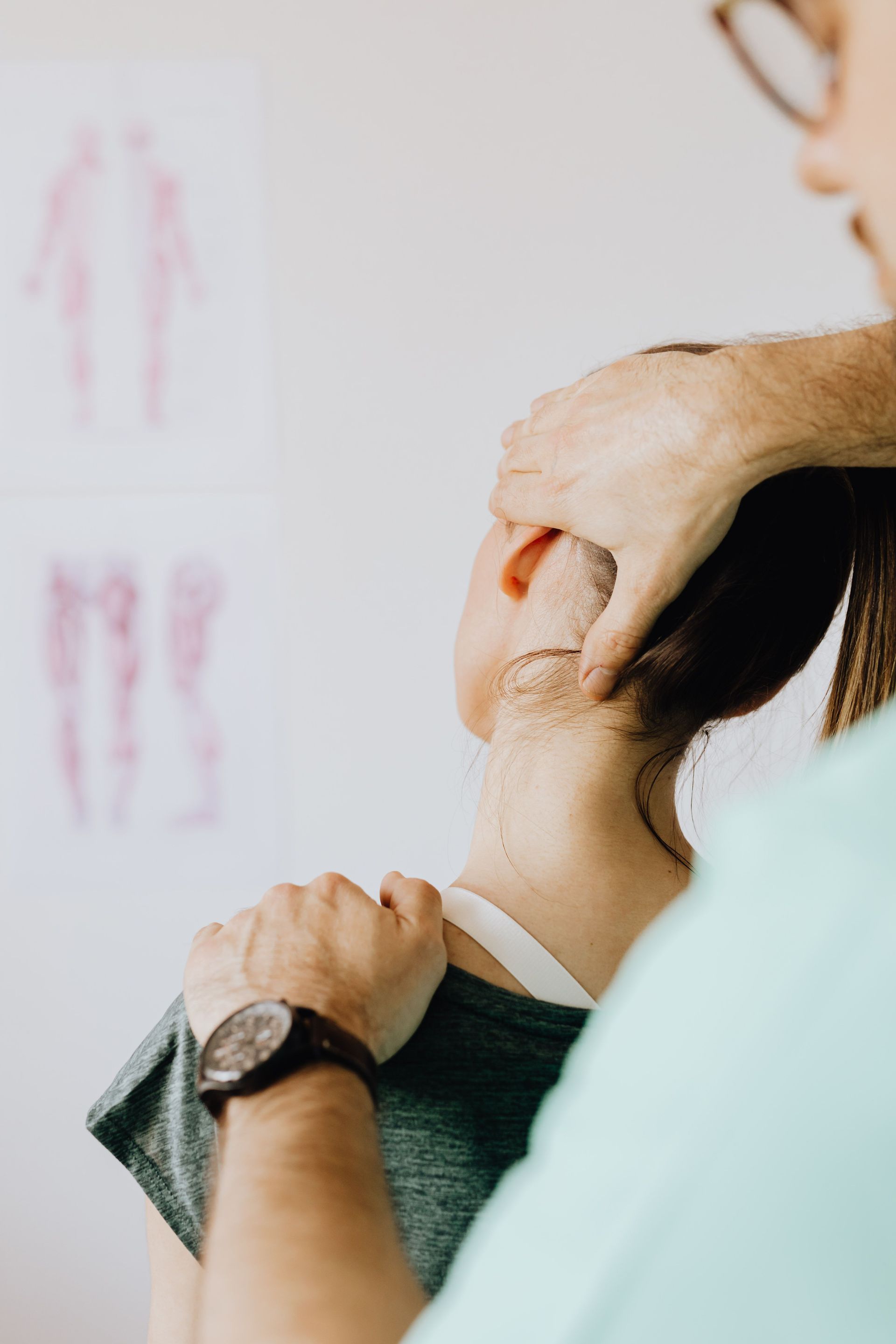 Person receiving a neck adjustment from a practitioner in a clinic, with anatomy chart in background
