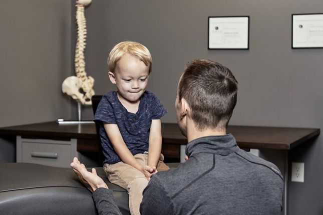 Doctor examining a smiling child in a clinic, with skeletal model and wall certificates in the background