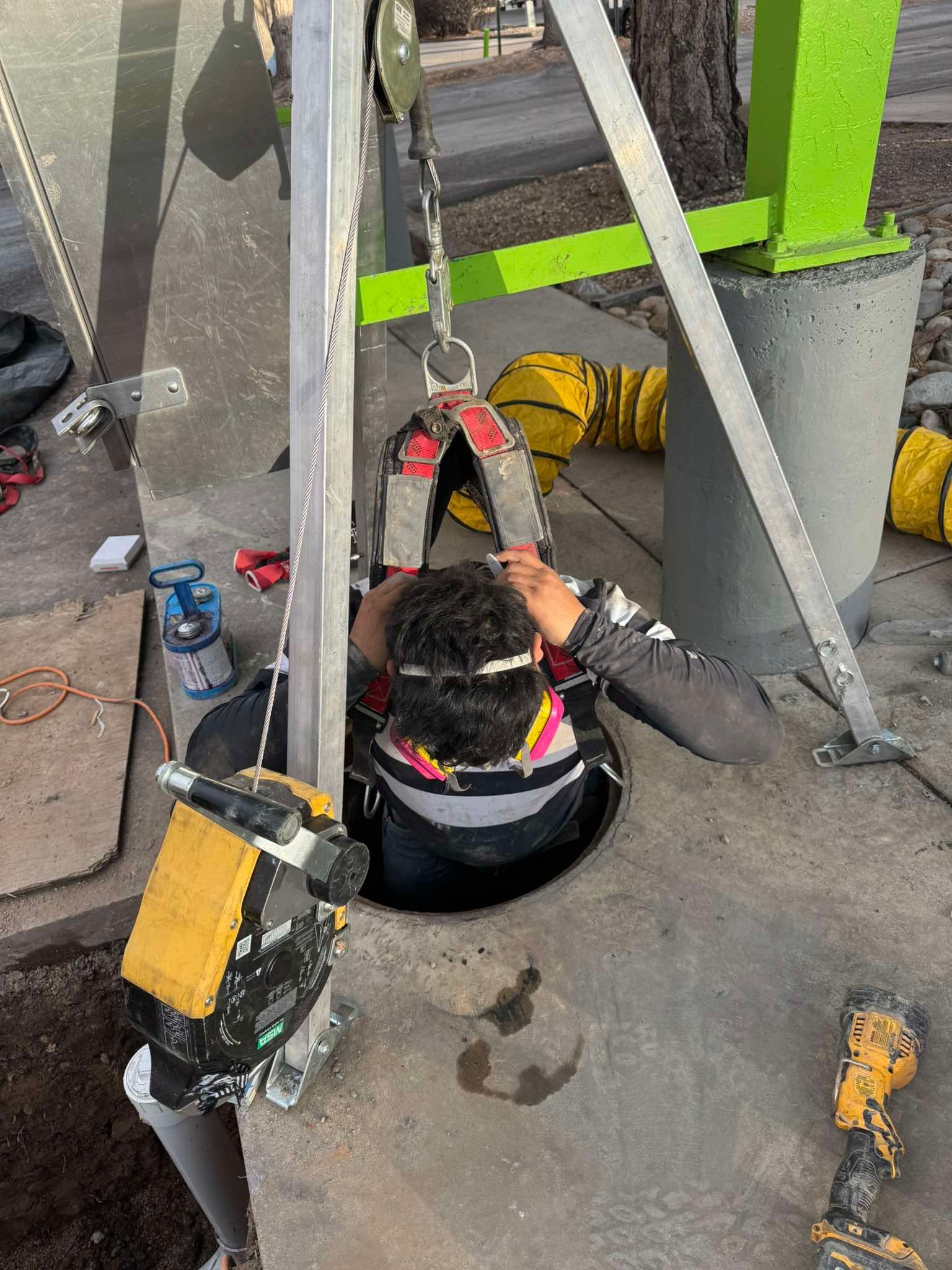 Worker in hard hat repairs equipment inside a concrete utility pit with tools and cables.