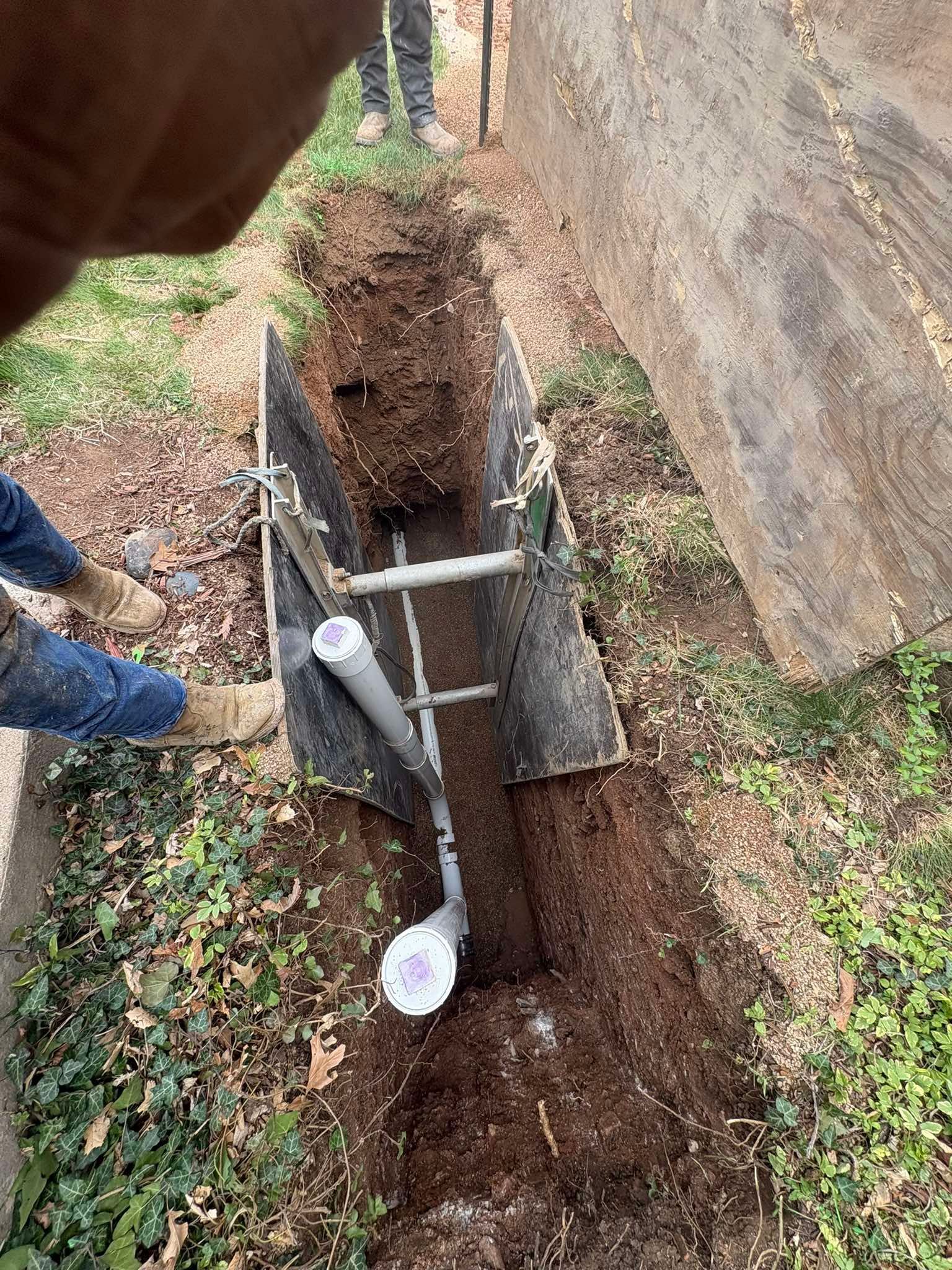 Person digging a narrow trench beside a wall, with a shovel and a temporary pipe or stake in the ground