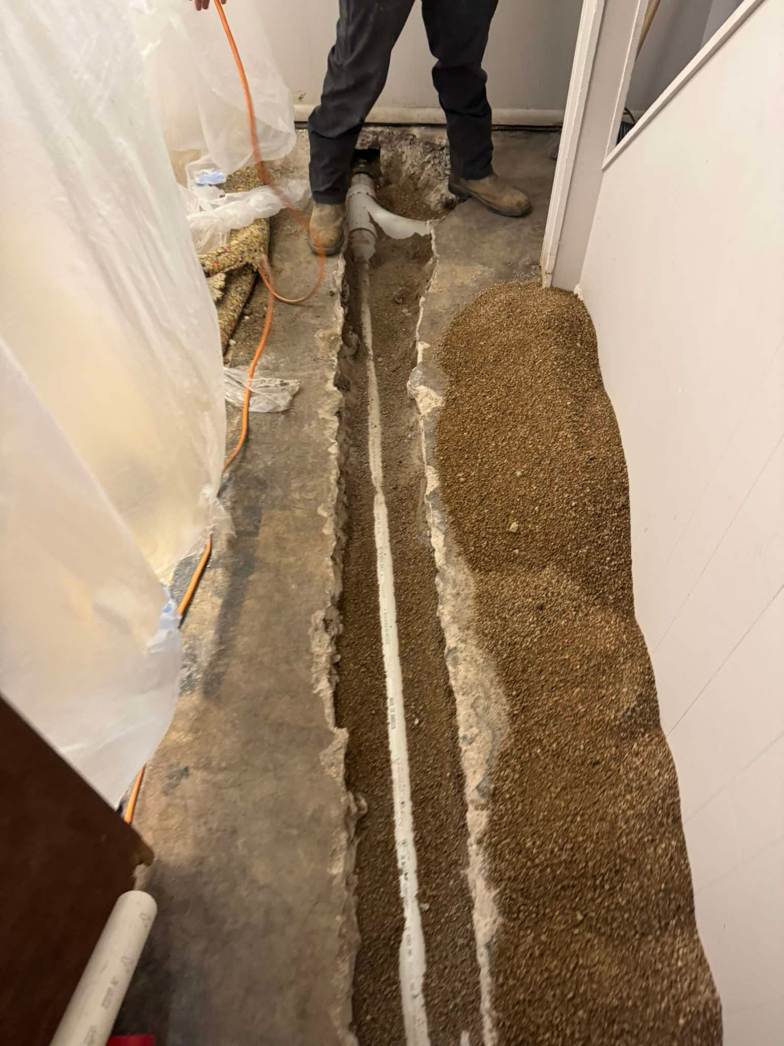Person standing in a narrow trench beside excavated soil and plastic sheeting during basement repair work