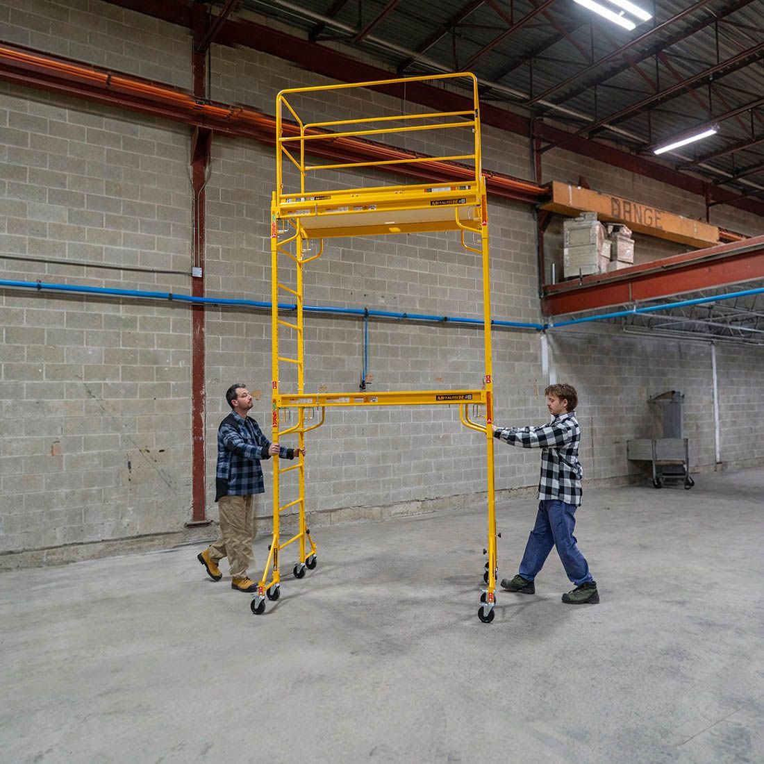 Dos trabajadores empujan un andamio móvil amarillo de gran altura en un espacio industrial similar a un almacén.