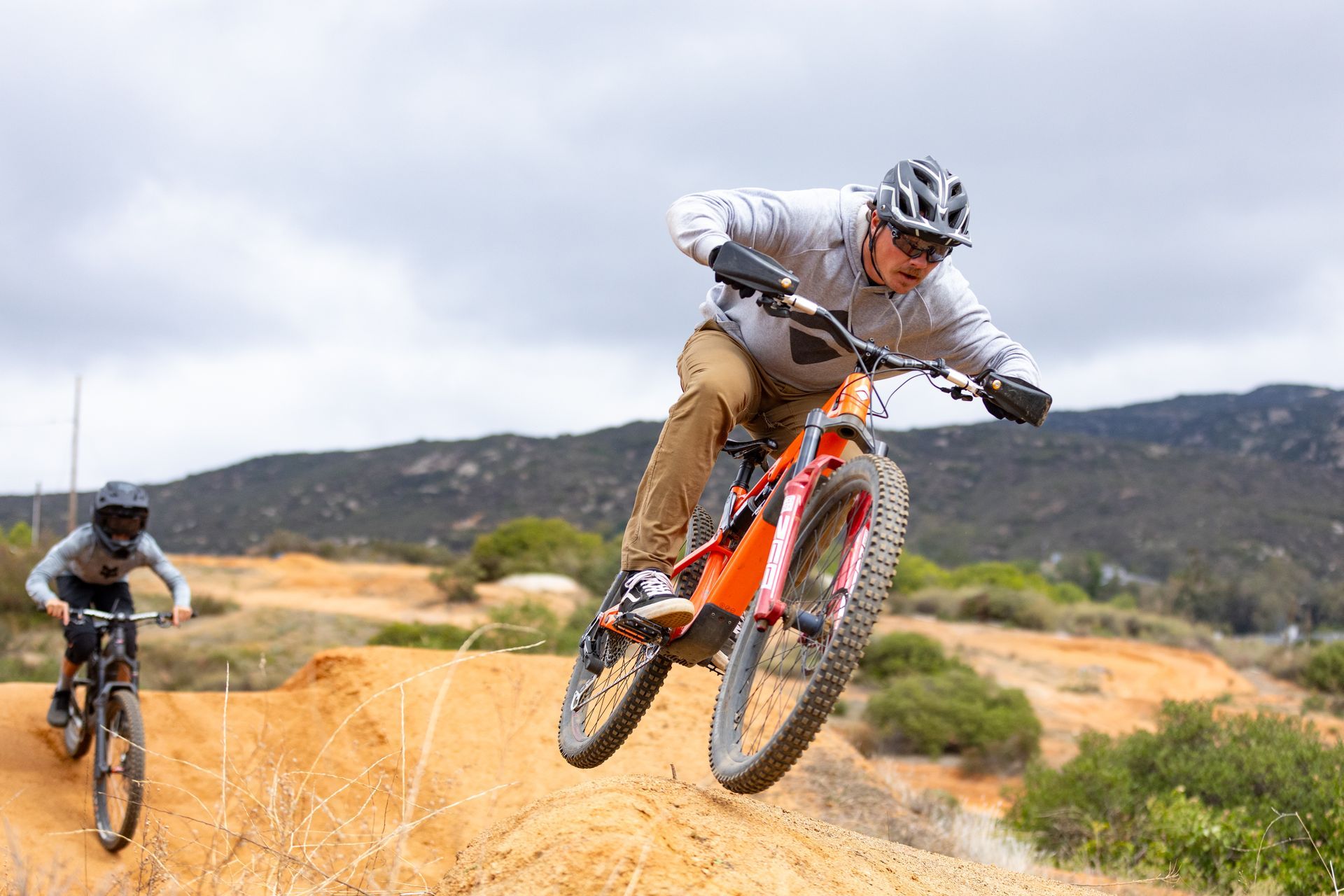 Two people on mountain bikes, one airborne on a dirt jump. The lead rider wears a helmet and is in mid-air