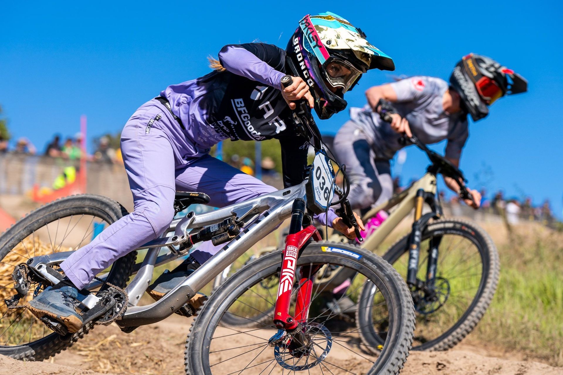 Two female mountain bikers racing downhill on a dirt track. One in lavender clothing, the other in gray. 