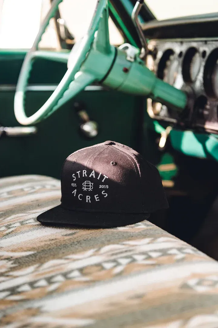 Black cap with white text rests on a patterned car seat, with a green steering wheel and dashboard in the background.