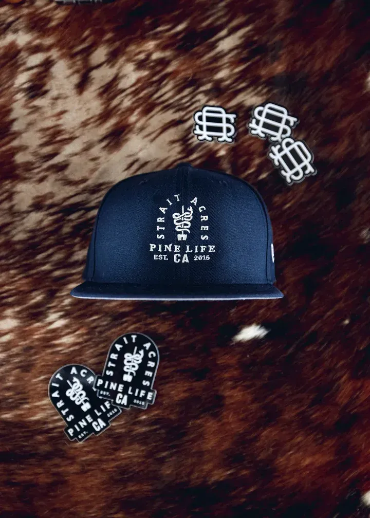 A navy blue Pine Life CA hat and enamel pins displayed on a brown hide background.
