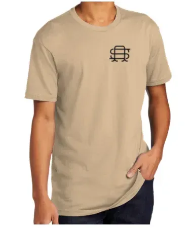 Tan t-shirt with a small black logo on the chest. A person wearing the shirt is standing with one hand in their pocket.