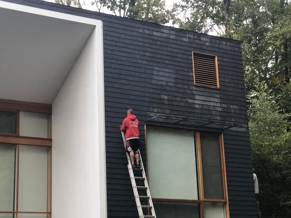 Person standing on a ladder painting a dark exterior wall beside large windows