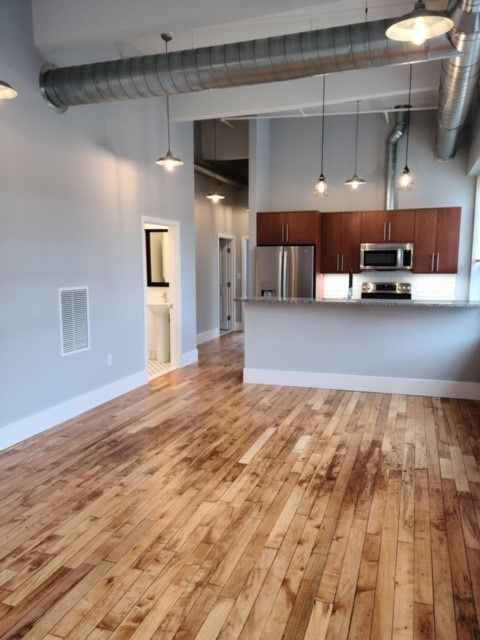 Empty loft apartment with hardwood floors, exposed ducts, and a kitchen with wood cabinets and stainless appliances