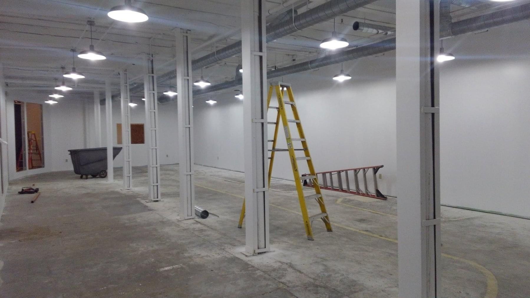 Empty unfinished industrial interior with white columns, exposed ceiling lights, and a yellow ladder.