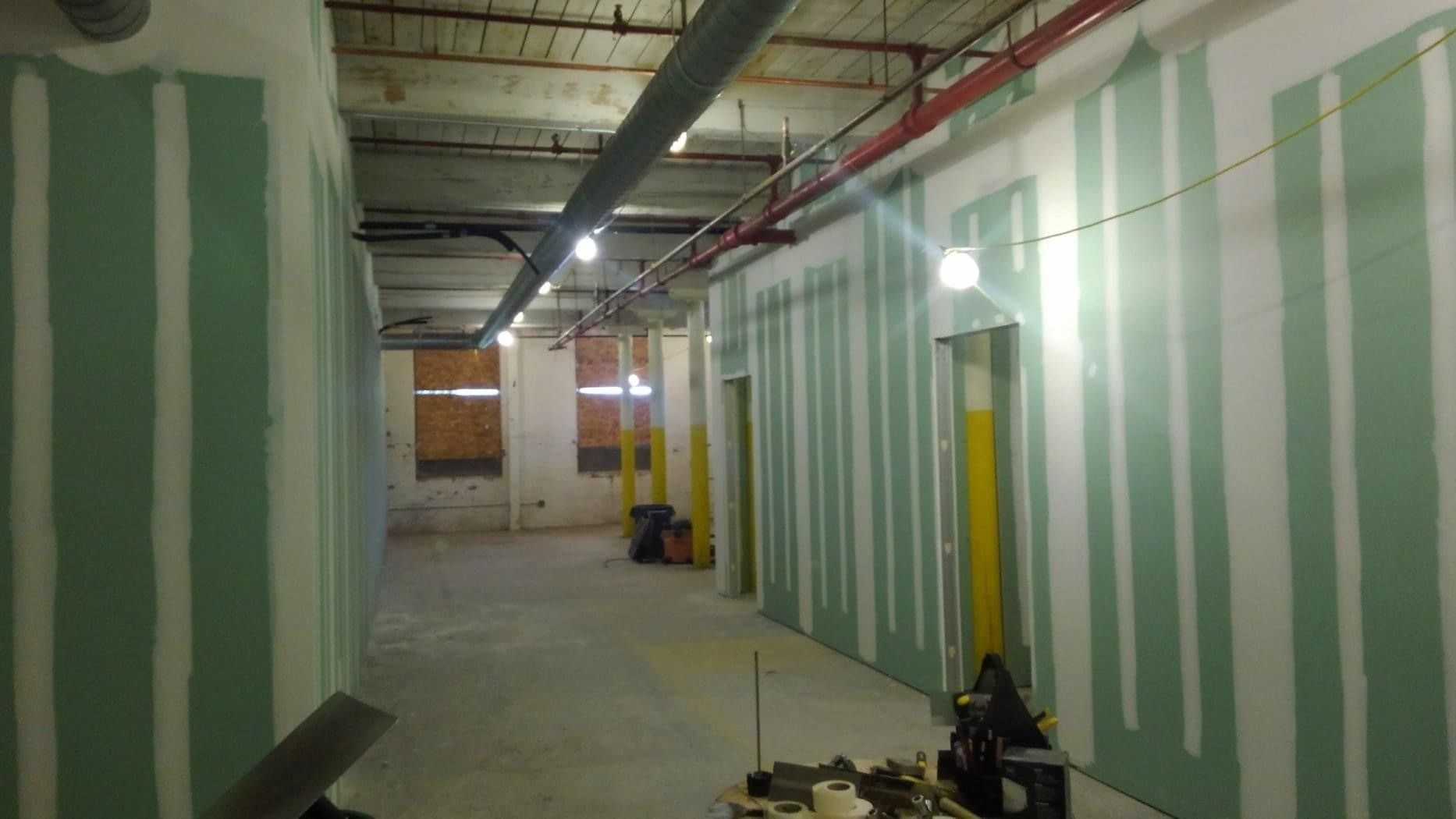 Unfinished hallway with green-striped drywall, exposed ceiling pipes, and construction materials on the floor