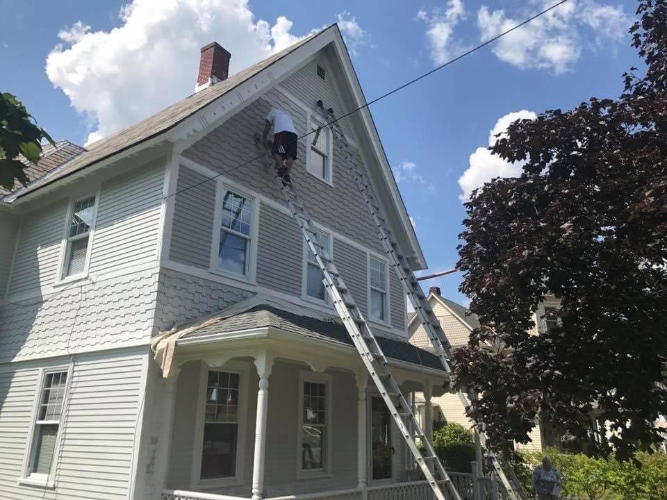 Two-story white house with a steep roof, porch, and ladder leaning against the side under a blue sky.