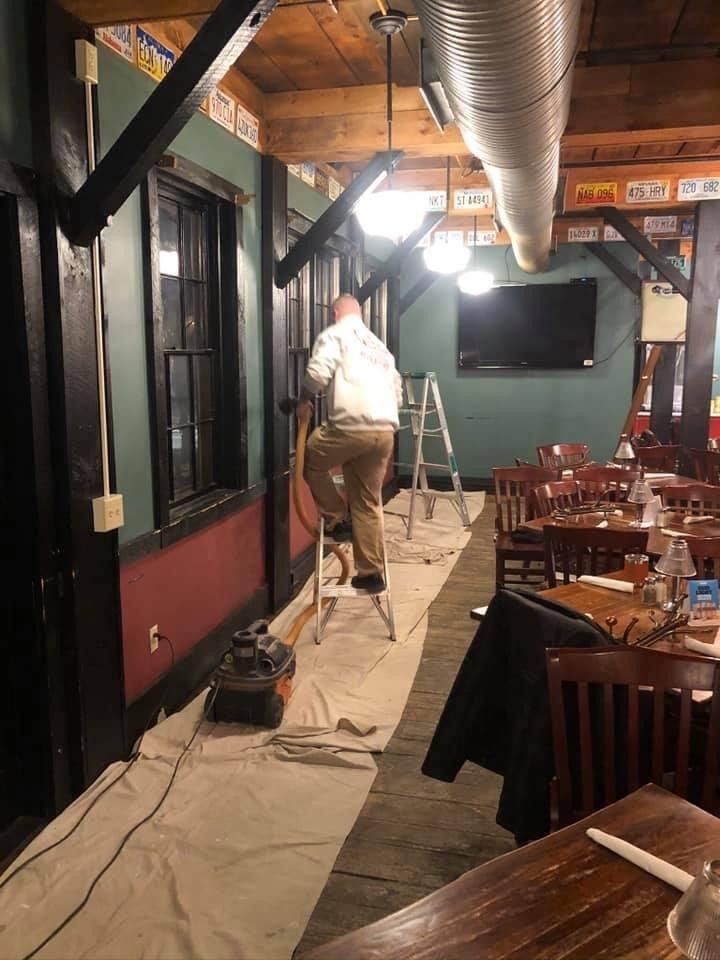 Worker painting a restaurant wall beside tables, chairs, and hanging ductwork at night.