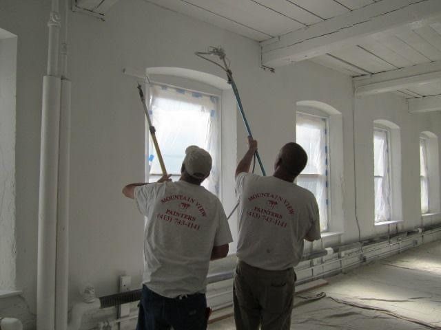 Two workers paint white interior walls in a bright room with tall windows.