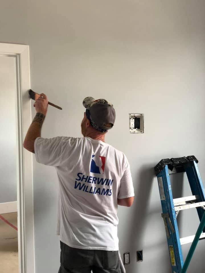 Painter in a white Sherwin-Williams shirt painting a wall inside a room, with a ladder nearby.