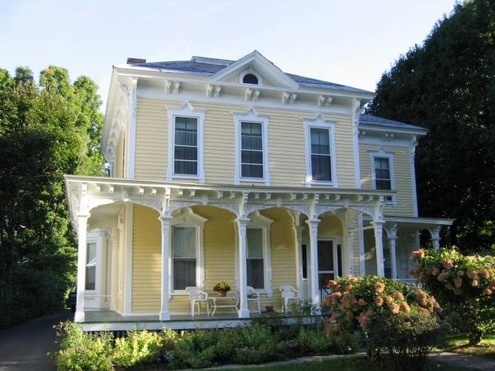 Yellow Victorian-style house with white trim and a wraparound porch, surrounded by trees and garden shrubs