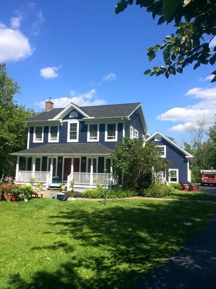Two-story dark blue house with white trim, front porch, and green lawn under a bright blue sky