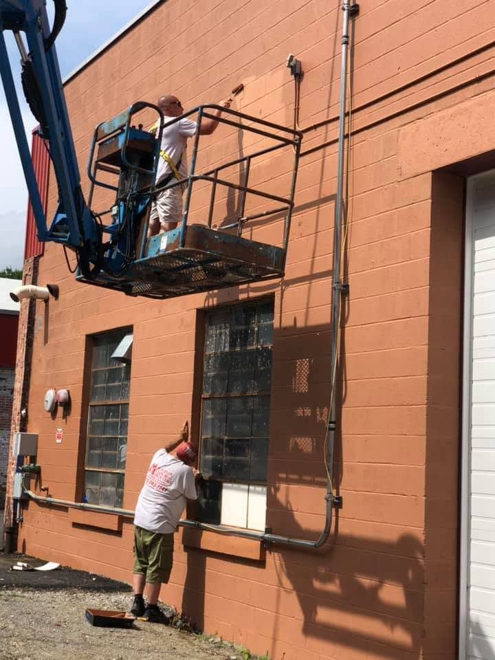 Worker on a blue lift painting a brick building while another person paints a lower window frame.