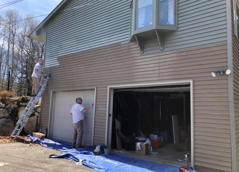 Workers painting a two-story house exterior beside open garage doors, one on a ladder and one below