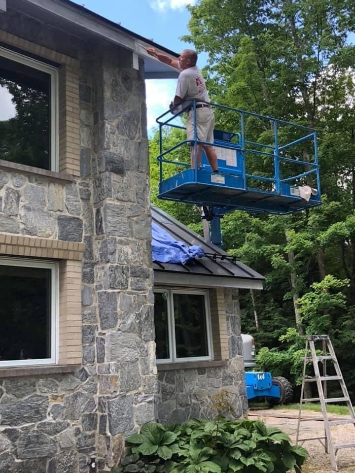 Worker on blue lift painting a stone house exterior beside ladders and trees