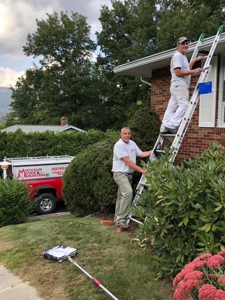 Two workers on a ladder painting a brick house exterior, with a red work van and lawn tools nearby.
