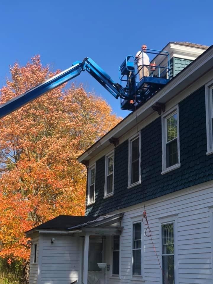 Worker in a blue lift repairs a roof beside a house with white siding and dark trim in autumn.