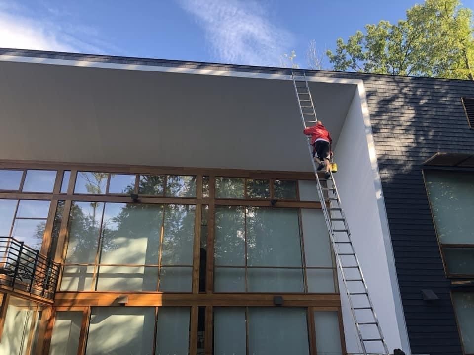 Person on a ladder cleaning a building facade under a gray overhang.
