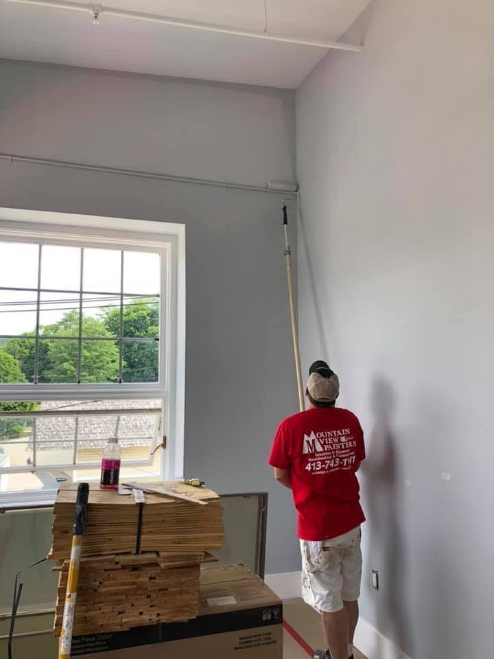 Person painting a white interior wall beside a window, standing on boxes in a room with stacked flooring boards.