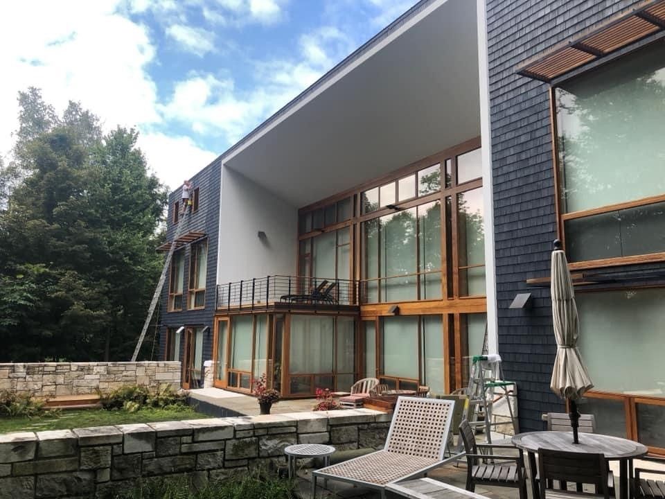 Modern house with large glass windows, balcony, and patio seating beside a small stone-lined lawn
