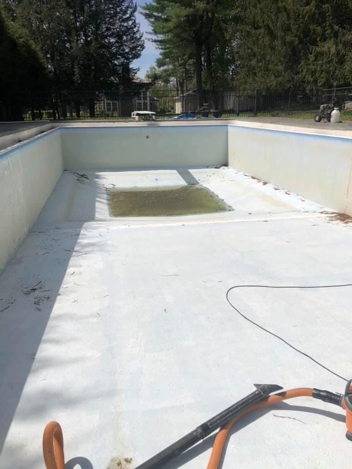 Empty drained swimming pool with white floor, a small green patch, and hoses in the foreground.
