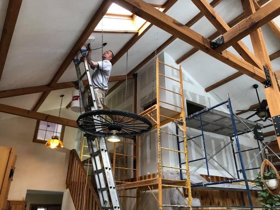 Worker on a ladder installing ceiling lights in a high-ceiling room with scaffolding and exposed beams