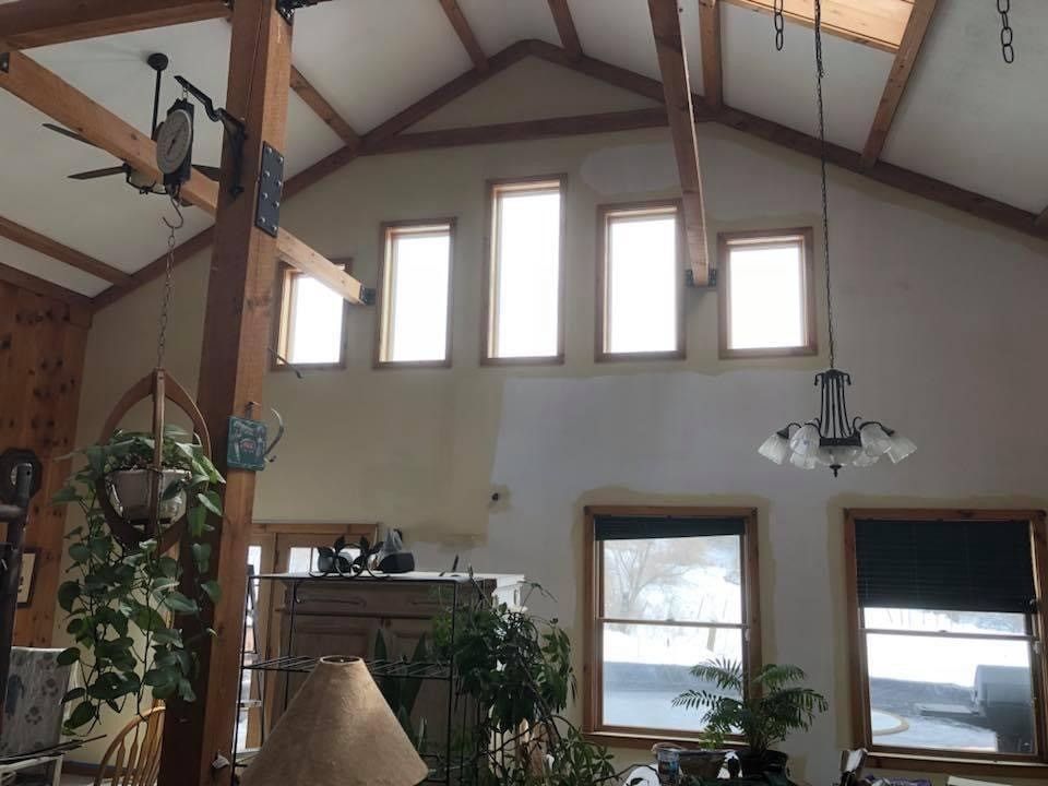 Cathedral-ceiling living room with tall windows, wood beams, hanging plants, and a view of water outside