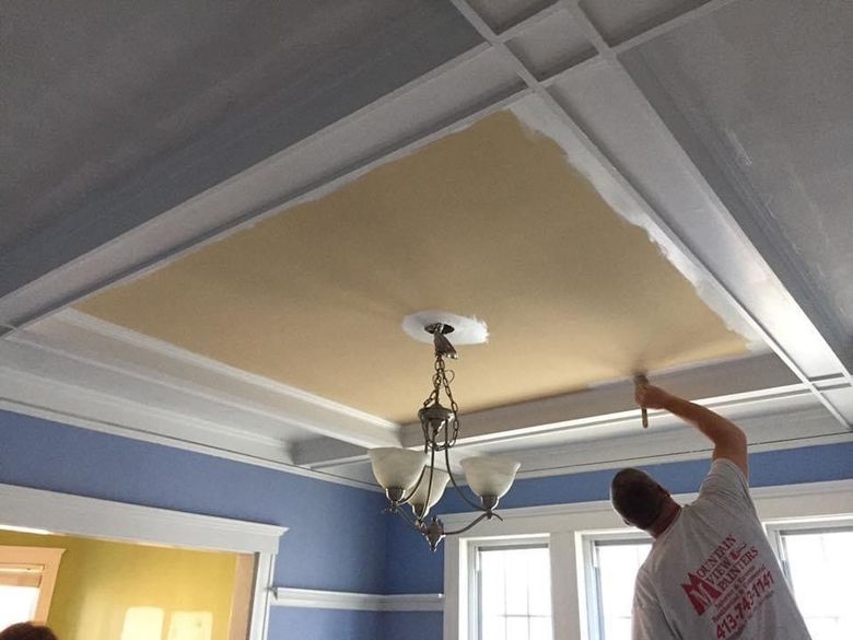 Person painting a beige ceiling in a blue room with a hanging light fixture and crown molding