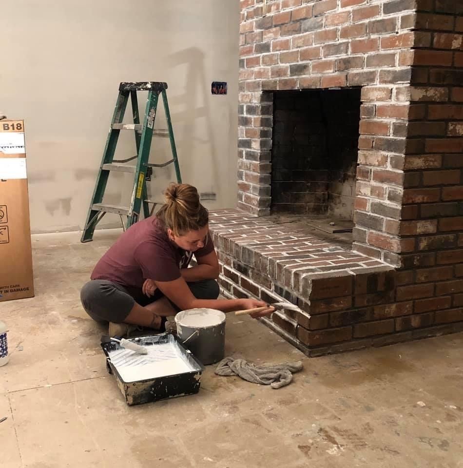 Person painting a brick fireplace in a room with a ladder and paint supplies