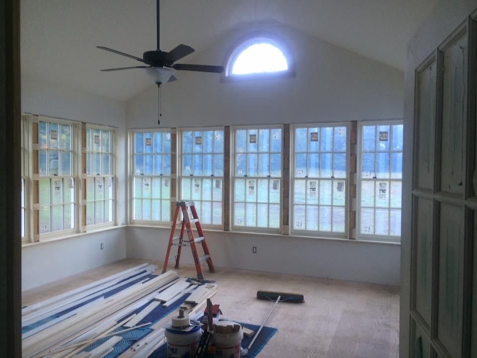 Bright sunroom under renovation with ladders, stacked boards, and windows along the wall