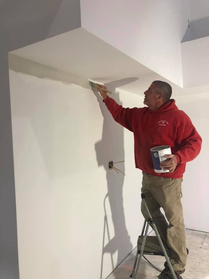 Painter in a red hoodie painting a white ceiling corner on a step stool.