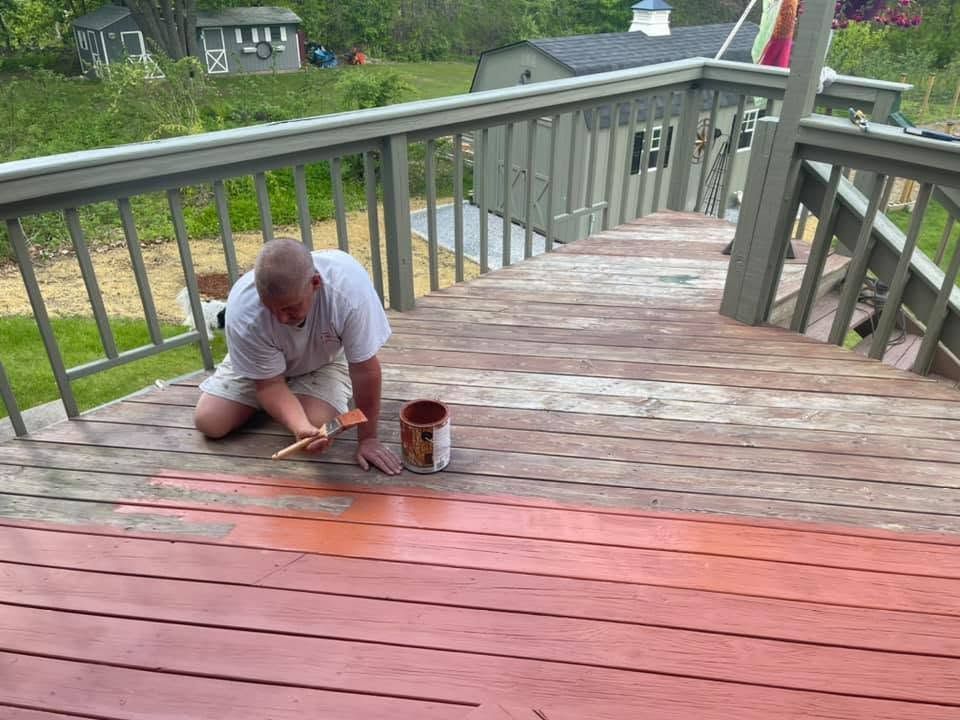 Man kneeling on a wooden deck, painting red planks near a paint can and railing
